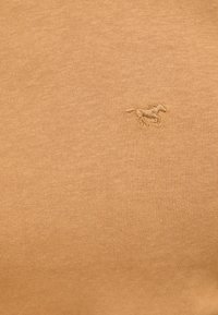 Hellbraunes Strickgewebe mit strukturierter Oberfläche. Verfügt über ein kleines, in etwas dunklerem Ton gesticktes Pferdelogo.