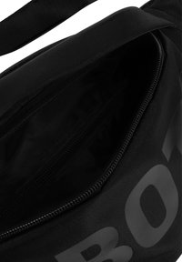 Czarna nylonowa torba z fakturowanym wykończeniem, z główną przegrodą na zamek oraz dużym nadrukowanym logo z przodu. Wnętrze jest jednolite, czarne.