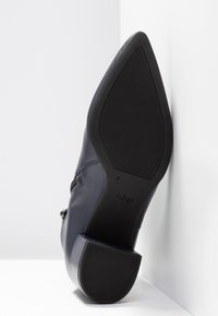 Bottine noire en cuir à bout pointu, présentant une semelle texturée et un talon bloc sur fond blanc.