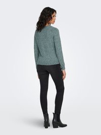 Roheline kootud kampsun ümmarguse kaelusega, sobitatuna mustade kitsaste teksade ja mustade kontsadega poolsaabastega. Tundub pehme ja tekstuuriga.