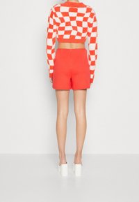 Suéter corto rojo con un patrón de cuadros blancos, combinado con pantalones cortos rojos de silueta ajustada y cremallera en la parte trasera. Zapatos de tacón bloque blanco.