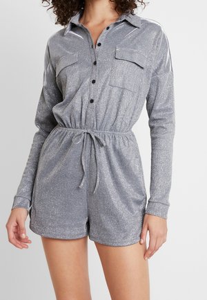 Žena v dlouhém střihu stříbrného třpytivého overal s předními kapsami, černými knoflíky a stahovací šňůrkou v pase na jednobarevném pozadí.