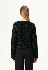 Veste noire avec texture florale en relief, manches longues et col rond. Associée à un pantalon noir bien taillé.