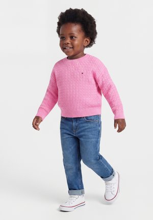 Tout-petit souriant qui marche, portant un pull rose en maille torsadée, un jean bleu retroussé et des baskets blanches sur un fond uni.