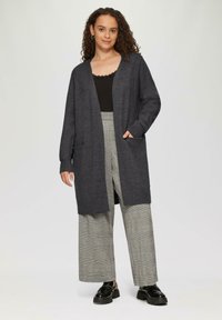 Długi szary kardigan z szalowym kołnierzem, dzianinowa tekstura, otwarty przód i dwie przednie kieszenie, zestawiony z szerokimi spodniami w kratę.