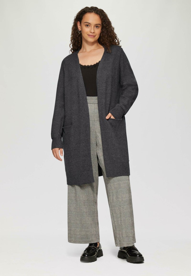 Długi szary kardigan z szalowym kołnierzem, dzianinowa tekstura, otwarty przód i dwie przednie kieszenie, zestawiony z szerokimi spodniami w kratę.