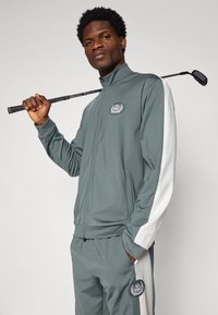 Siv športni trenirka z belimi stranskimi črtami, jakna na zadrgo in žepi. Ima okroglo oznako in drži golf palico.