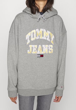 Personne portant un sweat à capuche gris avec le texte "TOMMY JEANS" en jaune et blanc, ainsi qu'un petit logo Tommy Hilfiger rouge, blanc et bleu sur la manche et la poitrine.