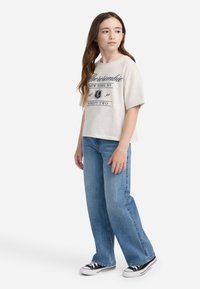 Bež majica s kratkim rukavima s mornarsko plavim grafičkim natpisom i grbom, uparena s svijetloplavim širokim trapericama i crnim platnenim tenisicama.