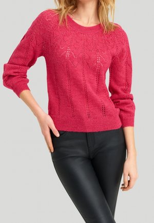 Mujer con un suéter rosa de punto con un patrón de hojas y pantalones de cuero negros, con la mano en la cadera y un fondo liso.