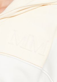 Krémová mikina s kapucňou s vyrazeným logom "MM" na hrudi, mäkkou textúrou a kontrastným prešívaním pozdĺž švov.