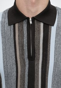 Gros plan sur une chemise tricotée avec des rayures verticales noires, grises et bleu clair, avec un col noir et une fermeture éclair frontale partiellement ouverte.