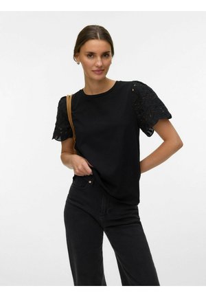 Kvinde iført sort broderet kortærmet top, sorte bukser og bærende en brun skuldertaske mod en ensfarvet hvid baggrund.