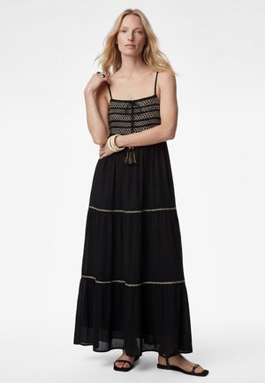 Femme aux longs cheveux blonds portant une robe maxi noire avec une broderie zigzag beige, des sandales et des bracelets blancs, debout devant un fond blanc.
