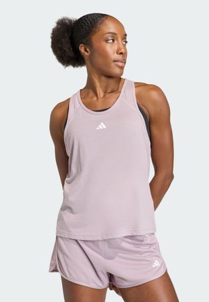 Atletische vrouw die een lichtpaarse Adidas tanktop en korte broek draagt, kijkt zijwaarts met haar handen achter haar rug, tegen een effen achtergrond.
