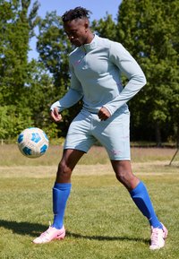 Light blue athletic tracksuit with pink accents, blue socks, and pink cleats. Player controls a patterned soccer ball on grass.