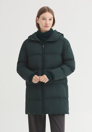 Femme debout portant une longue doudoune d'hiver vert foncé avec une capuche et un pull à col roulé sombre en dessous.