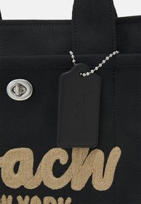 Musta kangaskassi, jossa on vaaleanbeige kirjailtu "Coach" -logo, hopeiset yksityiskohdat ja musta etiketti, joka on kiinnitetty hopealla palloketjulla.