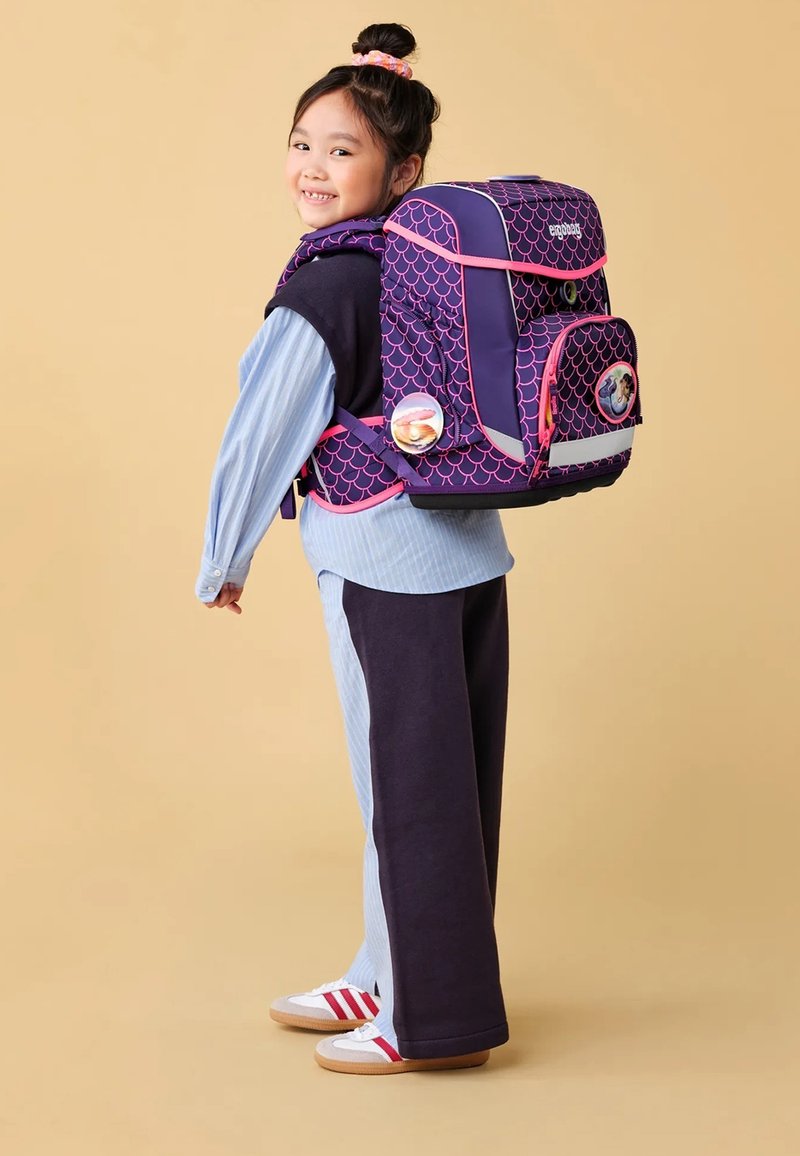 Mochila morada con diseño de escamas rosas, bolsillo frontal con cremallera, insignias laterales y correas acolchadas, llevada por un niño con ropa en capas.