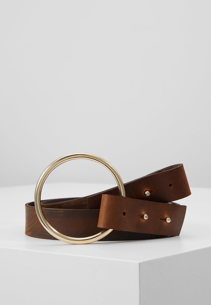 Ceinture en cuir marron enroulée avec une grande boucle ronde en métal doré et deux petites pointes métalliques, posée sur une surface blanche.