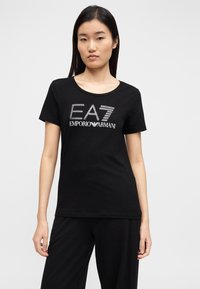 Μαύρο βαμβακερό κοντομάνικο t-shirt, με λογότυπο "EA7 EMPORIO ARMANI" με γκλίτερ στο μπροστινό μέρος, στρογγυλή λαιμόκοψη και κανονική εφαρμογή.