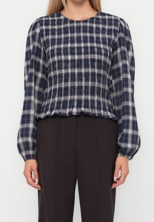 Femme portant une blouse à manches longues bleu marine et blanche à carreaux avec une taille froncée et un pantalon noir taille haute, debout devant un fond blanc.