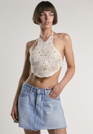 Kvinde med kort brunt hår iført en beige hæklet haltertop med perler og en lyseblå denimnederdel, poserer med den ene hånd i lommen.