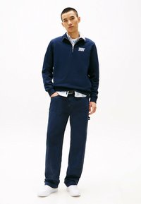 Marineblauwe sweatshirt met een halve rits en witte logo, gecombineerd met marineblauwe oversized broek en witte sneakers op een effen achtergrond.