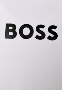 Vit t-shirt med fet, svart, versaler som stavar "BOSS" i mitten. Slät tygtextur med en minimalistisk design.