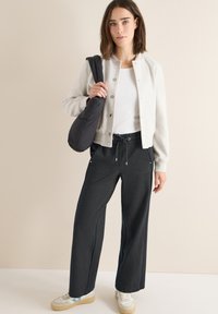 Lichtgrijze bomberjack over een wit shirt; zwarte joggingsbroek met zijpiping; witte sneakers met blauwe accenten; zachte texturen.