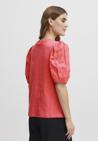 Tricou din bumbac corai cu mâneci scurte bombate, decorat cu broderie cu ochiuri. Croială lejeră, cu guler rotund, design minimalist.