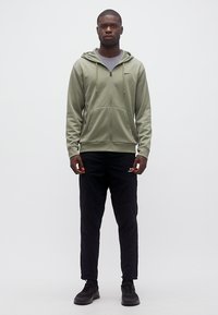 Άνδρας που στέκεται, φοράει ελιά φούτερ με φερμουάρ, γκρι μπλουζάκι, μαύρο παντελόνι Nike και μαύρα αθλητικά παπούτσια, μπροστά σε απλό λευκό φόντο.