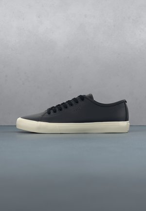 Niski czarny skórzany sneaker z czarnymi sznurowadłami i białą gumową podeszwą na niebieskiej powierzchni i szarym tle.