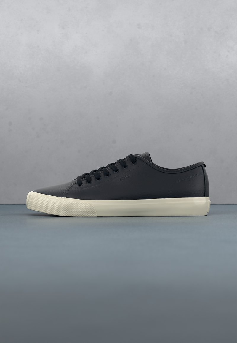 Baskets en cuir noir à profil bas avec lacets noirs et semelle en caoutchouc blanche sur surface bleue et fond gris.