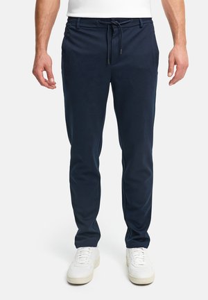 Man in marineblauwe slim-fit broek met trekkoorden in de taille en witte sneakers, staand tegen een effen achtergrond.