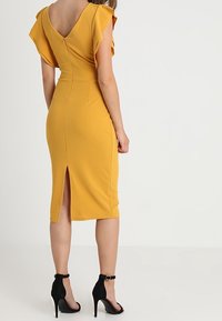 Senapsgul klänning med korta fjärilsärmar, figursydd kropp, knälång med en slits bak. Bärs med svarta högklackade skor.