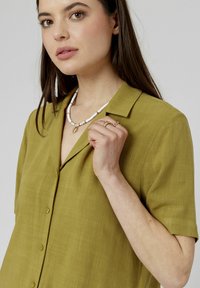 Chemise à col olive vert avec des manches courtes, fermeture à boutons sur le devant, tissu texturé, portée avec un collier blanc et doré.