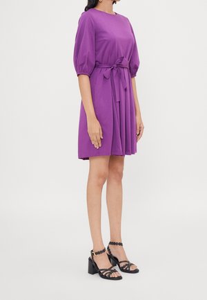 Robe violette à manches courtes avec un corsage ajusté et une jupe évasée, dotée d'une ceinture à la taille. Associée à des sandales à talons noires.