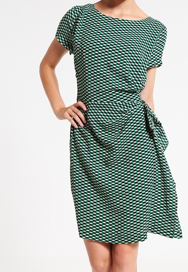 Femme portant une robe à manches courtes avec un motif géométrique vert et noir, un col rond et un détail noué sur le côté.