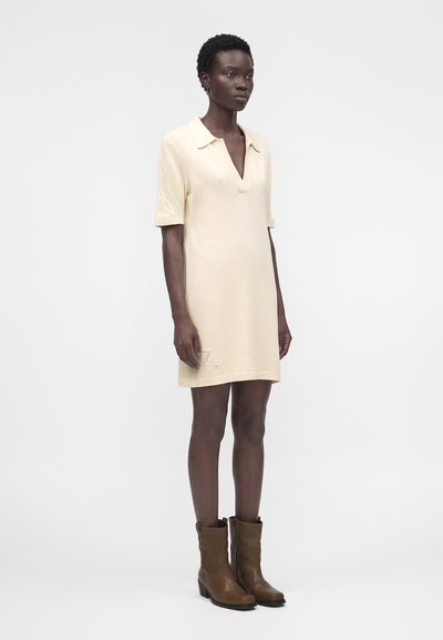 Γυναίκα με κρεμ κοντομάνικο φόρεμα με γιακά και καφέ μπότες μέχρι τη μέση της γάμπας, στέκεται μπροστά σε λευκό φόντο.