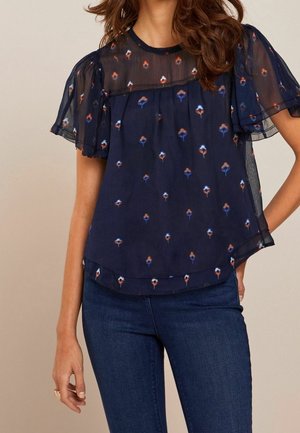 Femme portant un chemisier transparent bleu marine à imprimé floral et manches volantées, associé à un jean bleu foncé sur un fond beige.