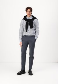 Overhemd met knopen en bloemmotief, grijze broek, en zwarte trui over de schouders gedrapeerd. Zwarte schoenen met een gestructureerde zool.