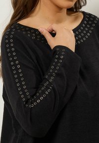 Pull noir avec des œillets le long de l'encolure et des manches. Texture douce avec une coupe décontractée, mettant en valeur les œillets.