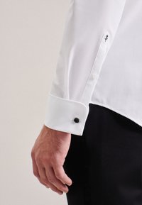 Camicetta bianca con bottone nero, polsini singoli con un piccolo ricamo di rosa nera. Tessuto liscio, vestibilità su misura, design elegante.