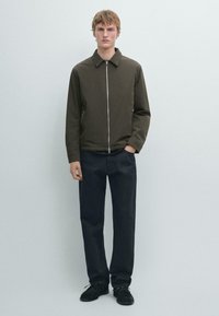 Ung mand iført en mørkeolivengrøn lynlåsjakke, sorte bukser og sorte sko, stående foran en ensfarvet lysegrå baggrund.