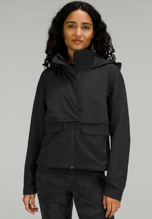 Frau trägt eine schwarze Kapuzenjacke mit großen Klappentaschen und schwarze Hose mit Camouflage-Muster, steht vor einem einfarbigen grauen Hintergrund.