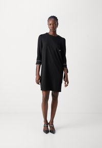 Black dress with three-quarter sleeves, round neckline, and white piping on cuffs. Smooth texture, knee-length, worn with black heeled shoes.