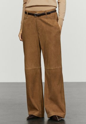 Person trägt breite braune Wildlederhose mit schwarzem Gürtel, beiges langärmeliges Oberteil und dunkle spitze Schuhe, steht auf grauem Boden.