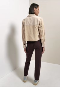 Zkrácená béžová kordurová bunda s texturovaným vzorem a knoflíkovými manžetami, spárovaná s tmavě hnědými kalhotami a světlými teniskami.