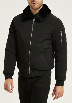 Homme portant une veste bomber noire avec col en fourrure, fermeture éclair à l'avant, poignets côtelés et poche zippée sur la manche gauche.